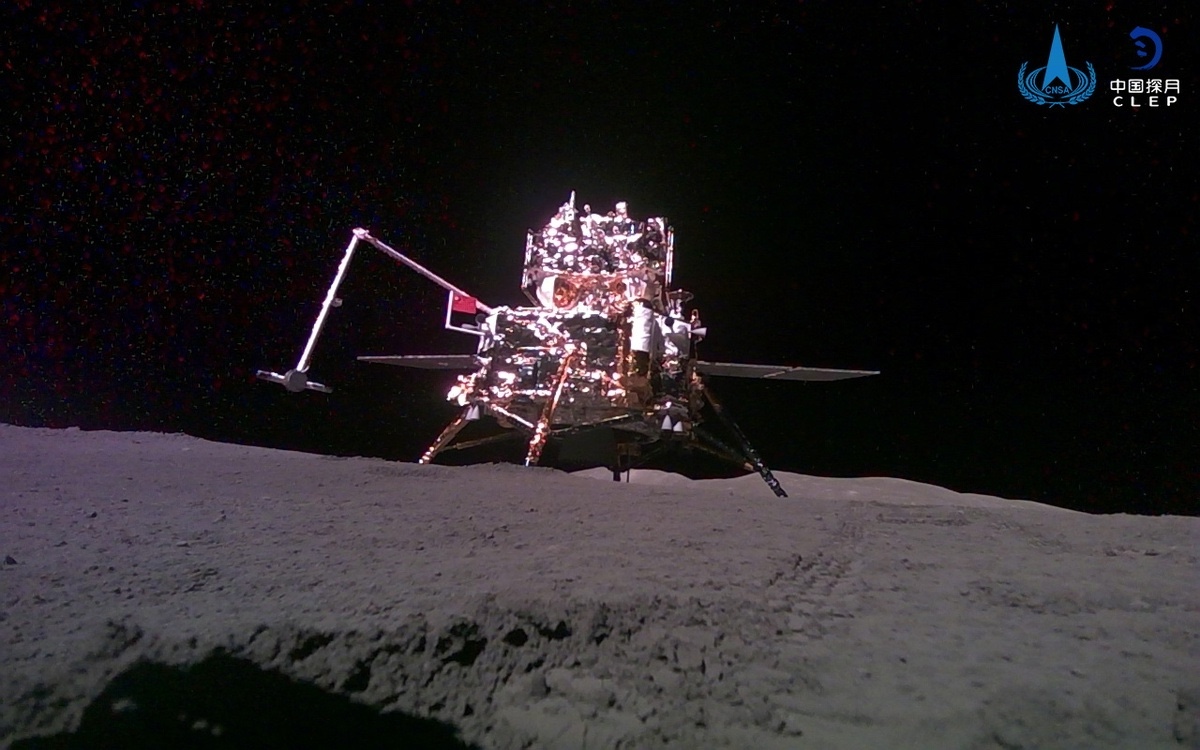 A lunar lander sits on the moon's surface with solar panels extended, under a black sky with stars. A logo is visible in the upper right corner.