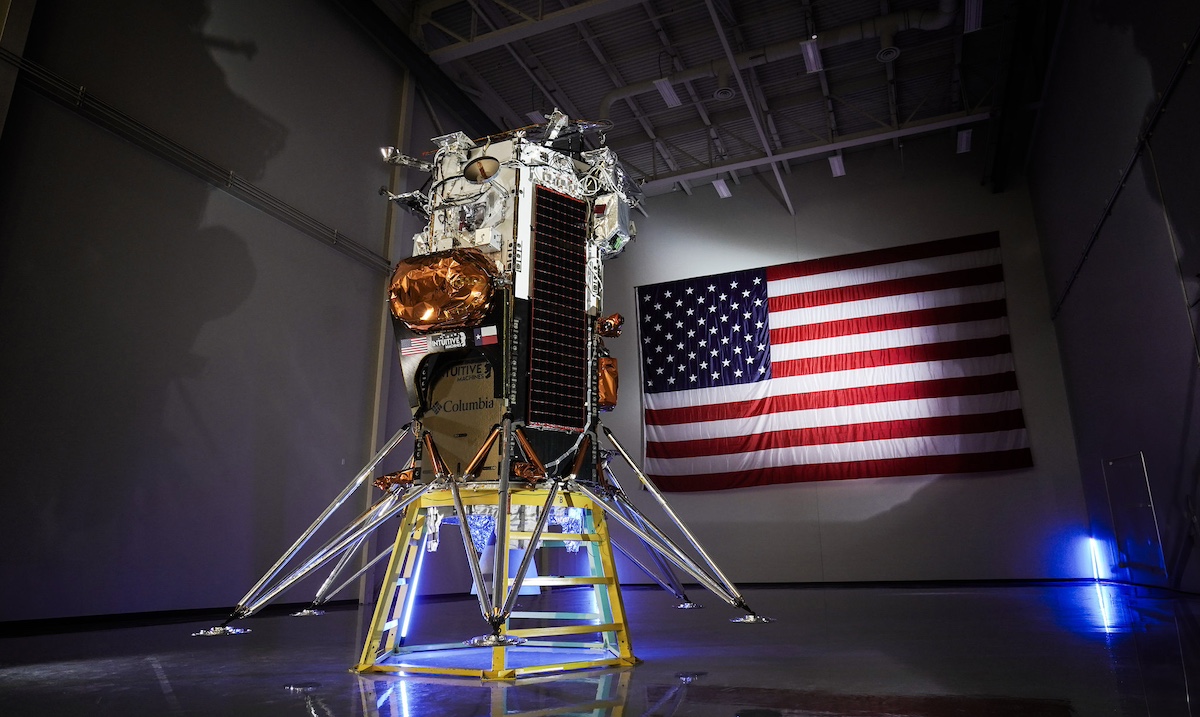 A spacecraft lander displayed indoors with a large American flag on the wall behind it.