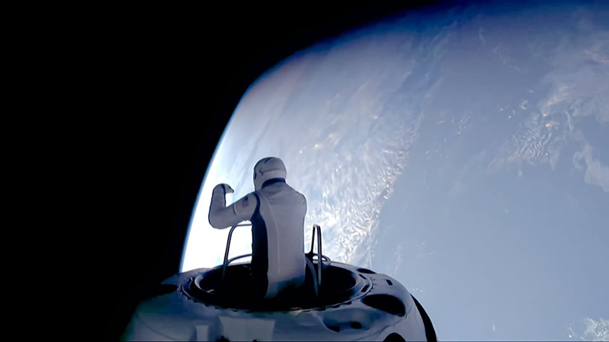 An astronaut in a spacesuit stands in an open hatch, looking out at Earth from space.