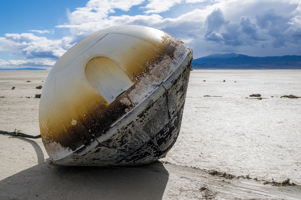 A scorched, round spacecraft lies on a barren, sandy surface under a cloudy sky, with mountains in the background.