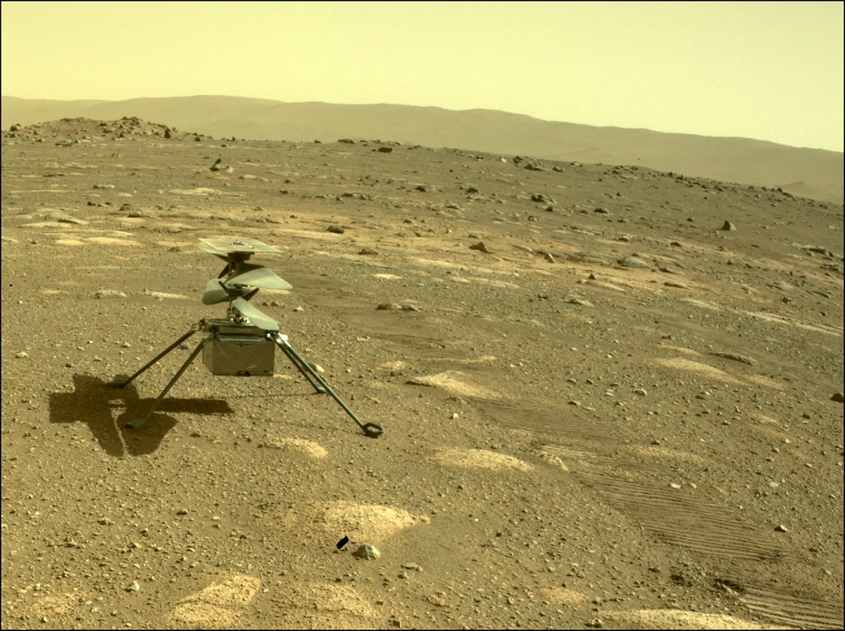 A small robotic helicopter sits on a rocky, dusty landscape under a pale sky on Mars. There are hills visible in the background.