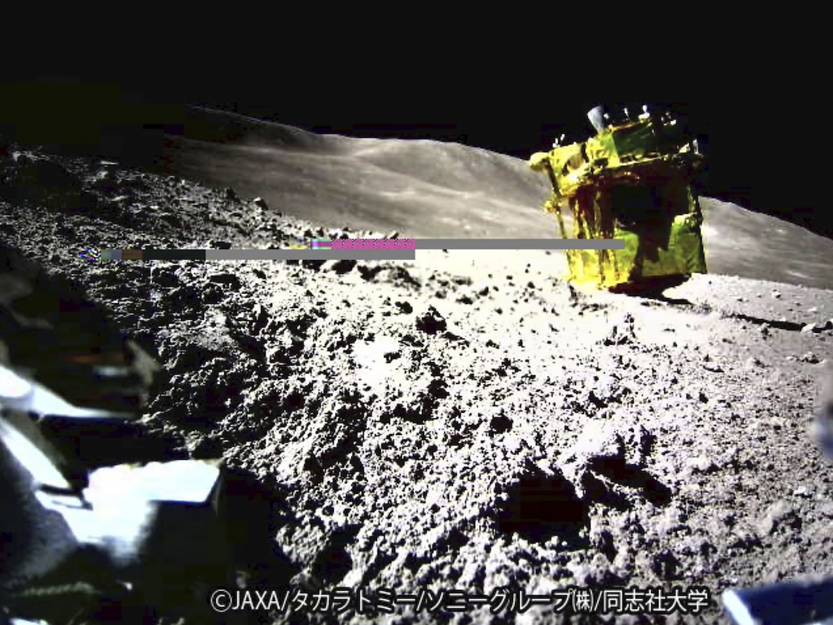 A yellow spacecraft, possibly the Hayabusa2 lander, is perched on a rocky surface of an asteroid, with rough terrain visible under a clear sky. Visible text includes ©JAXA and Japanese characters.