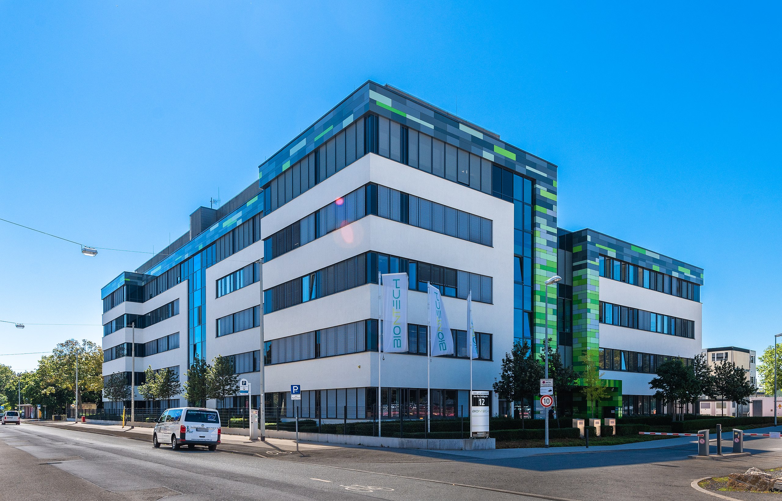 The modern multi-story office building, with its white and glass facade featuring green accents, stands prominently. The street in front, lined with trees and a few parked vehicles, provides a breath of fresh air—a stark contrast to the looming threat of lung cancer in urban environments.