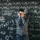 A person stands in front of a chalkboard filled with complex mathematical formulas, covering their face with their hands.