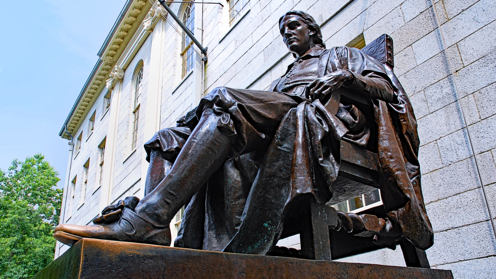 A bronze statue of a seated man in historical attire, symbolizing the global elite, is prominently positioned in front of a stately stone building.