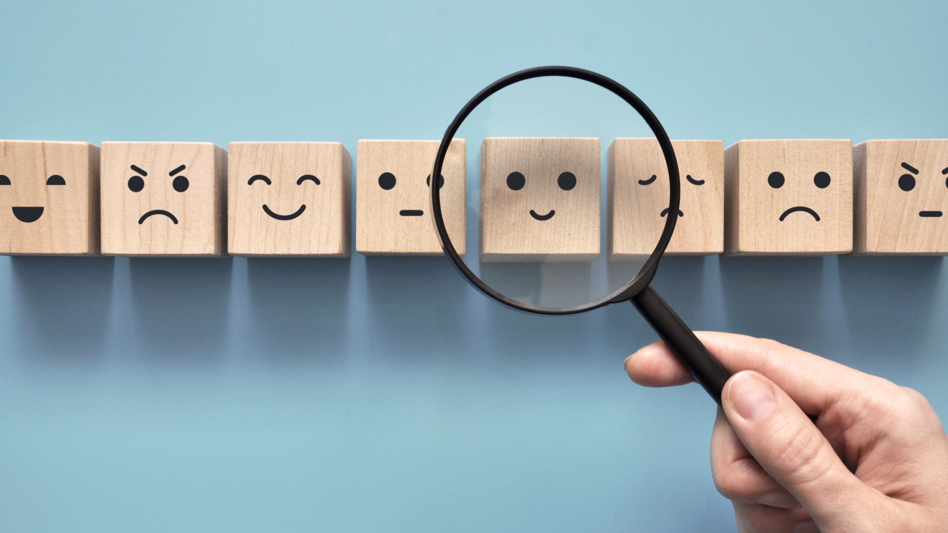 A hand holds a magnifying glass over wooden blocks with various facial expressions, focusing on a happy face against a blue background.