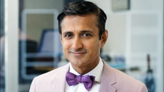 A man in a light pink suit and purple bow tie smiles slightly, standing indoors with blurred office background.