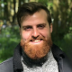 A smiling person with a red beard stands outdoors in a wooded area.