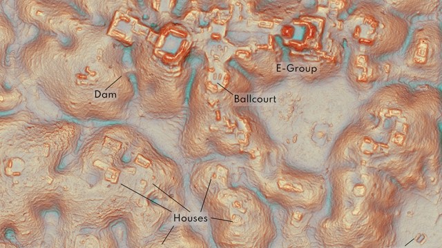 An aerial view of a topographic map showing labeled features: Dam, Ballcourt, E-Group, and Houses, with varying elevation and terrain details.