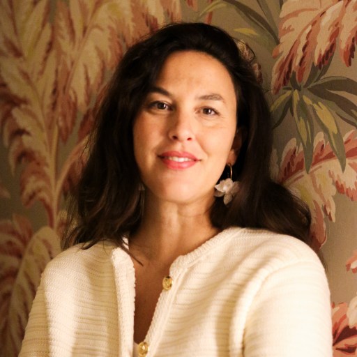 A person with long dark hair smiles, wearing a light-colored top and earrings, standing against a patterned wallpaper with leaf designs.