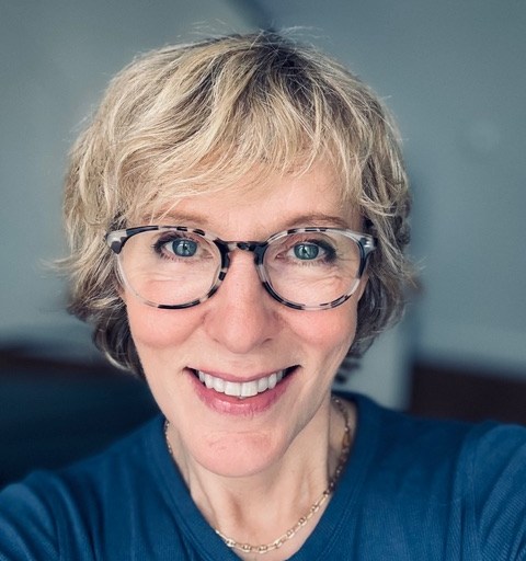 A person with short blond hair and glasses smiles at the camera, wearing a blue top and a necklace.