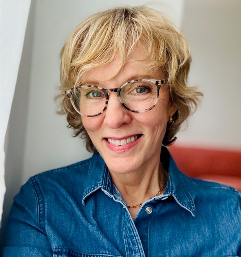 A woman with short blonde hair and glasses smiles while wearing a denim shirt.