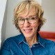 A woman with short blonde hair and glasses smiles while wearing a denim shirt.