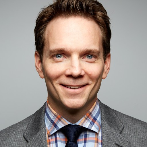 A man in a gray suit and checkered shirt with a dark tie smiles at the camera against a plain background.