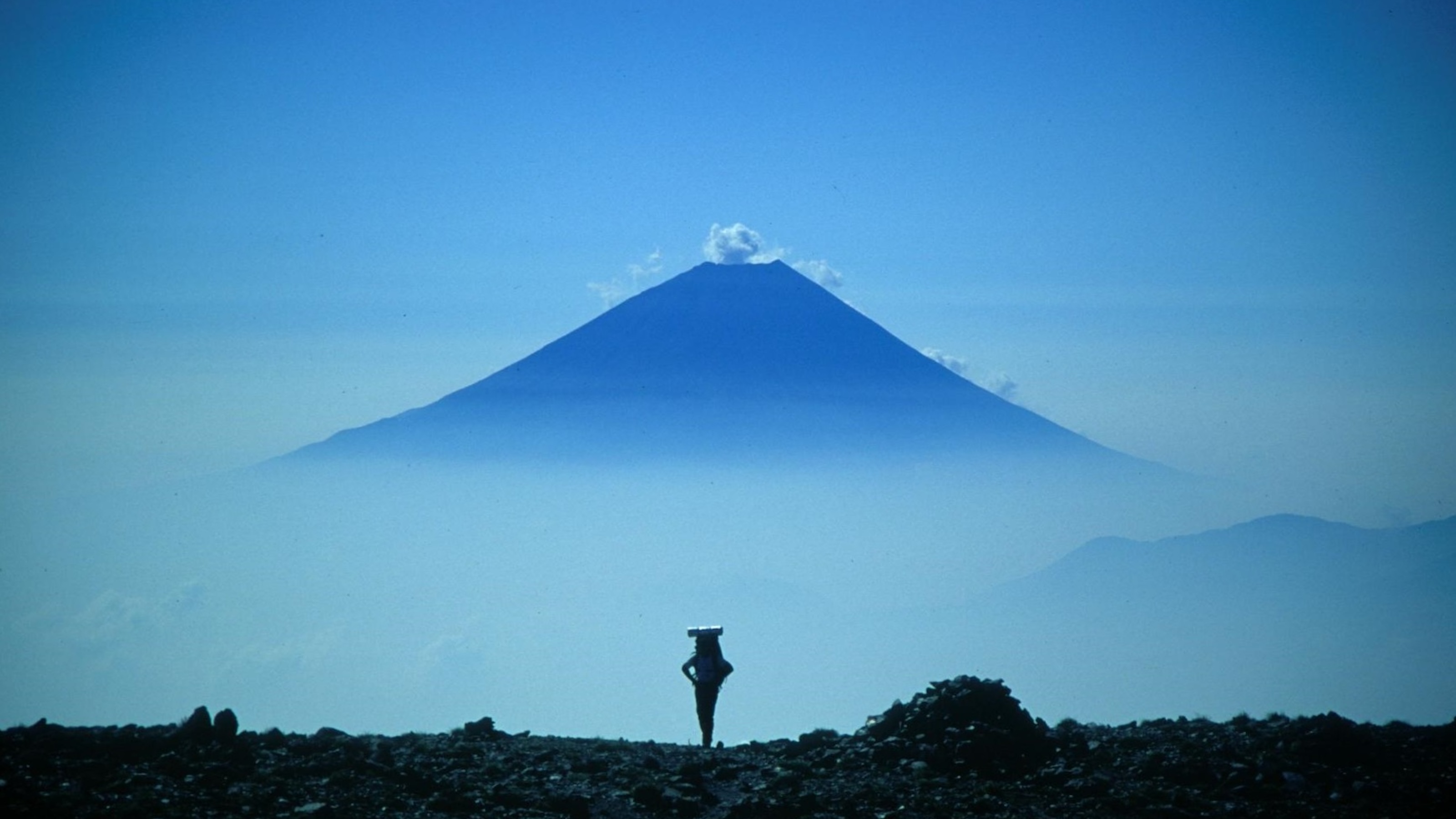 A person with a backpack stands on rocky terrain, contemplating the distant, mist-covered volcanic mountain under a clear blue sky, pondering the longevity of nature's breathtaking beauty.