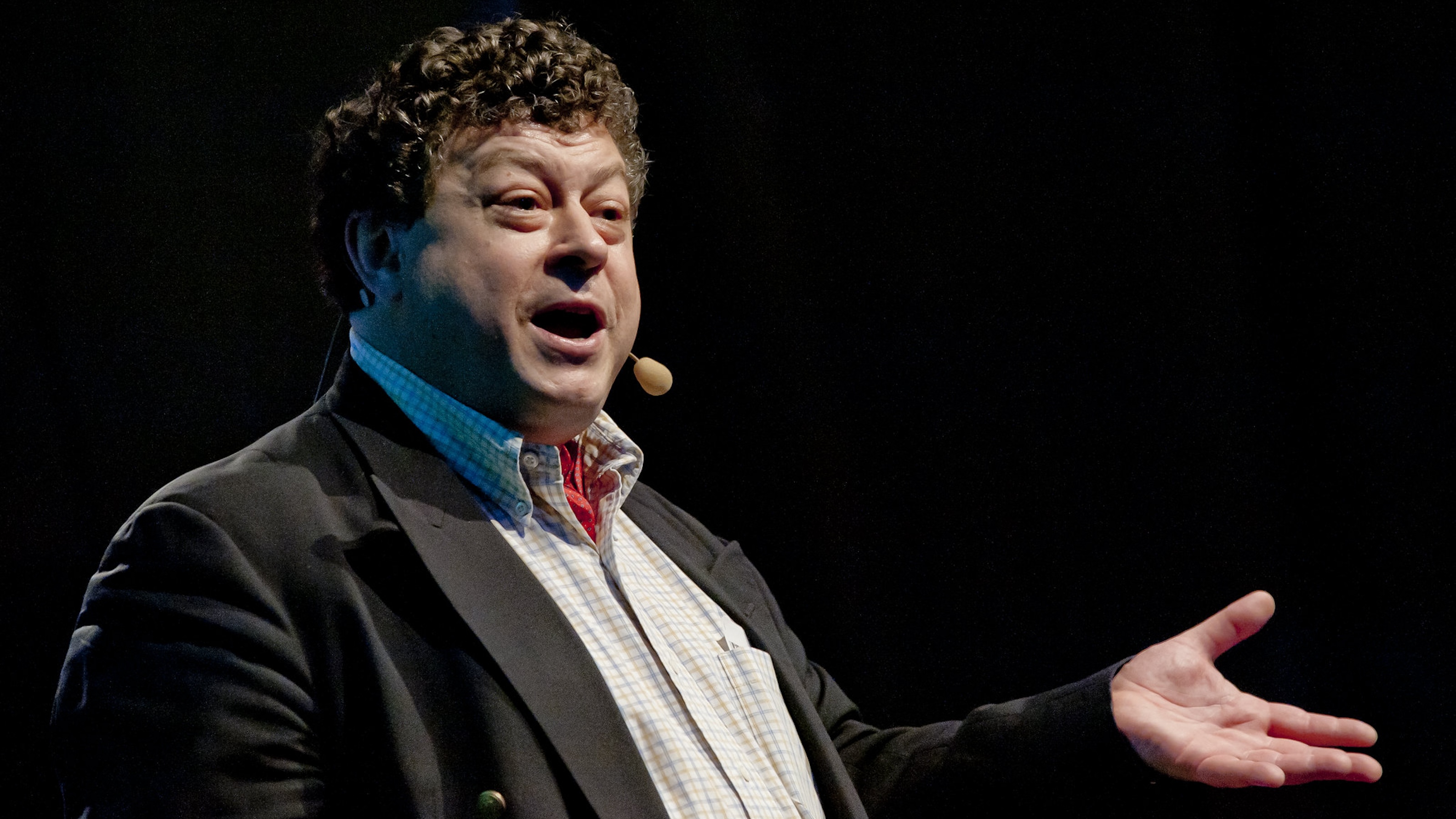 A man with curly hair wearing a suit and headset microphone gestures while speaking on stage.