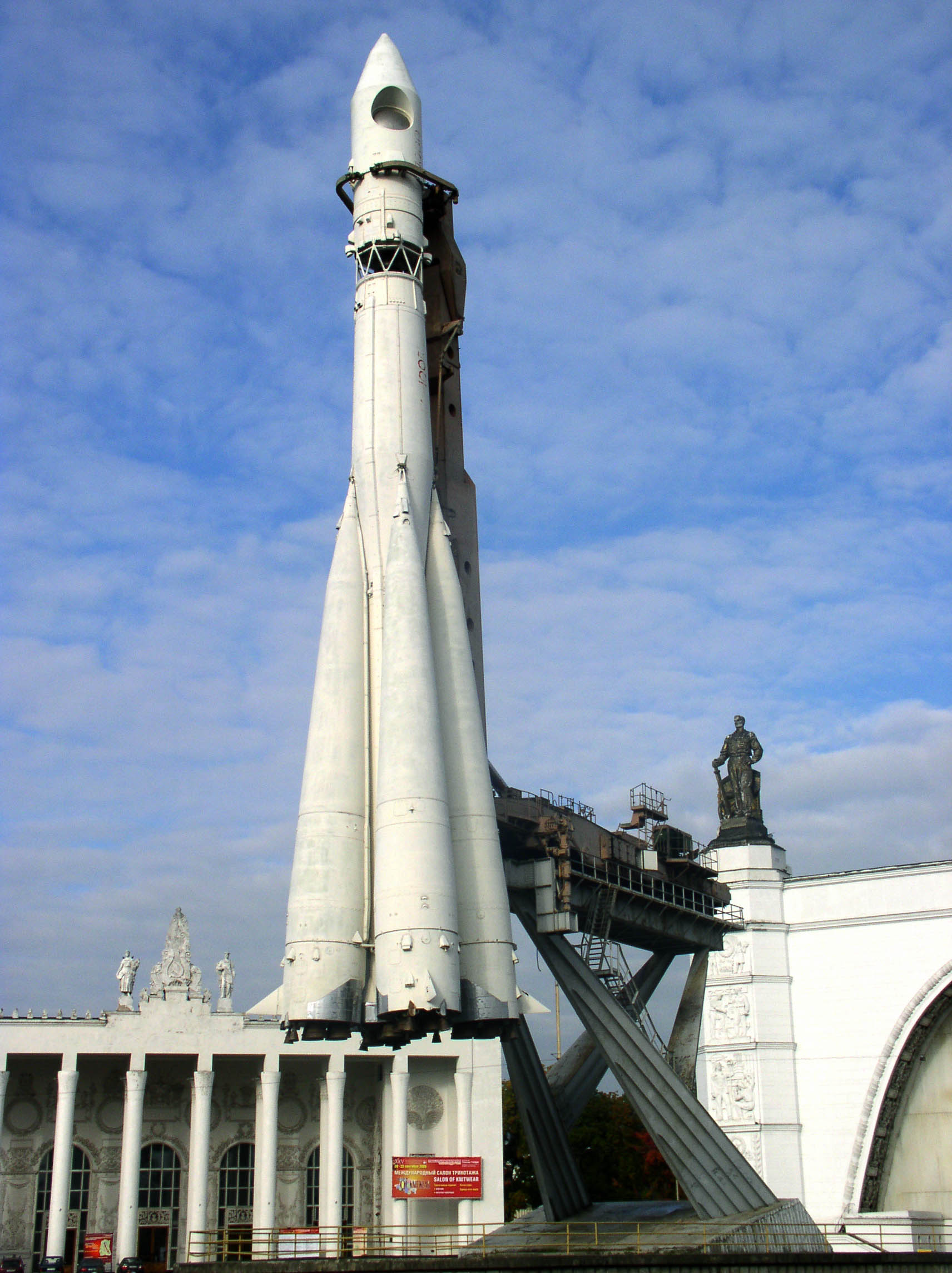 A tall white rocket, reminiscent of the USA's lunar missions, stands vertically on a launchpad against a cloudy blue sky. Nearby is a white building and a statue, evoking the legacy of the space race against the USSR.