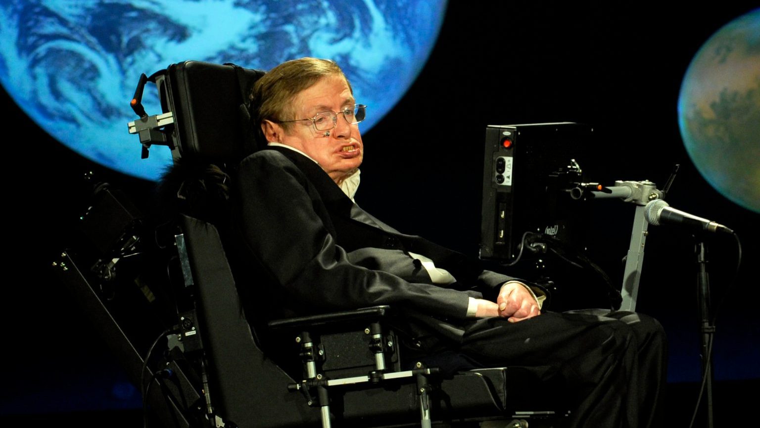 A person in a motorized wheelchair speaks into a microphone, their voice reminiscent of Stephen Hawking's, as stunning earth images are projected in the background.