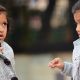 Two children in gray coats are standing outdoors. One is gesturing with her hand while the other looks on attentively.