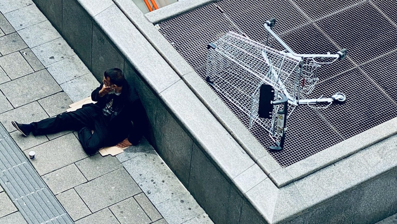 A person sits on a sidewalk near an escalator entrance, next to an overturned shopping cart on a metal grate.