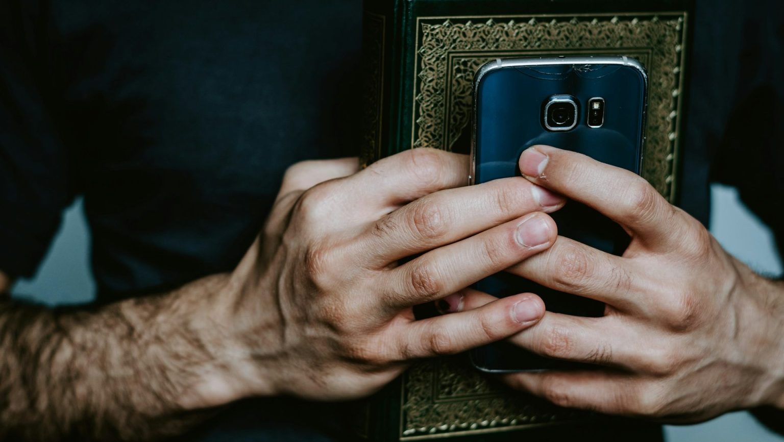 A person holds a smartphone and a book with an ornate cover, clasped together in both hands.