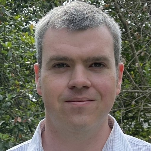 A person with short gray hair stands outdoors, wearing a patterned collared shirt. Green foliage is visible in the background.