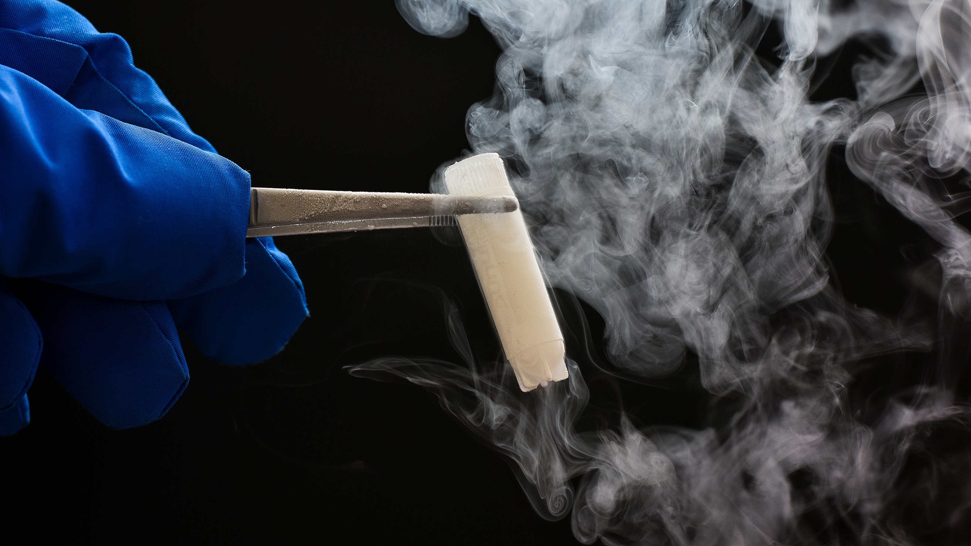 A gloved hand holds a smoking test tube with tongs against a dark background.