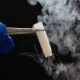 A gloved hand holds a smoking test tube with tongs against a dark background.