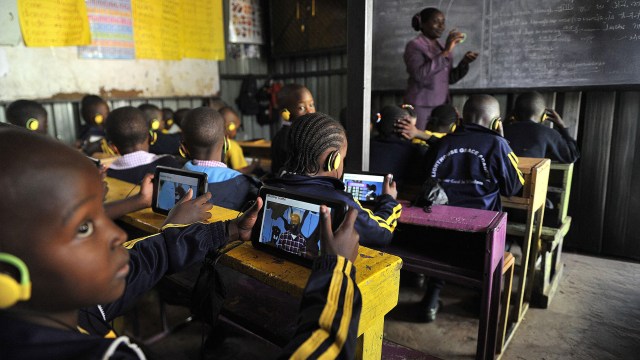 Children in a classroom use tablets with headphones while a teacher writes on a chalkboard.