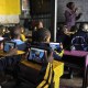 Children in a classroom use tablets with headphones while a teacher writes on a chalkboard.