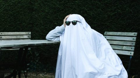 Person disguised as a ghost with sunglasses, sitting at an outdoor table.