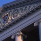 A close-up of a neoclassical building's facade with ornate carvings and columns, featuring an emblem at the center, reflects the grandeur often associated with the global elite.