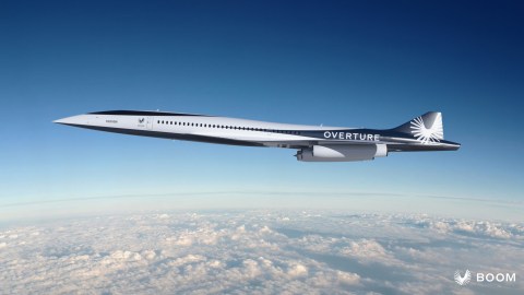 A sleek supersonic jet labeled "Overture" by Boom flies above the clouds against a clear blue sky.