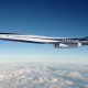 A sleek supersonic jet labeled "Overture" by Boom flies above the clouds against a clear blue sky.