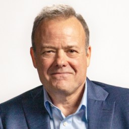 Ron Garan in a blue suit jacket and light blue shirt is smiling against a plain background.