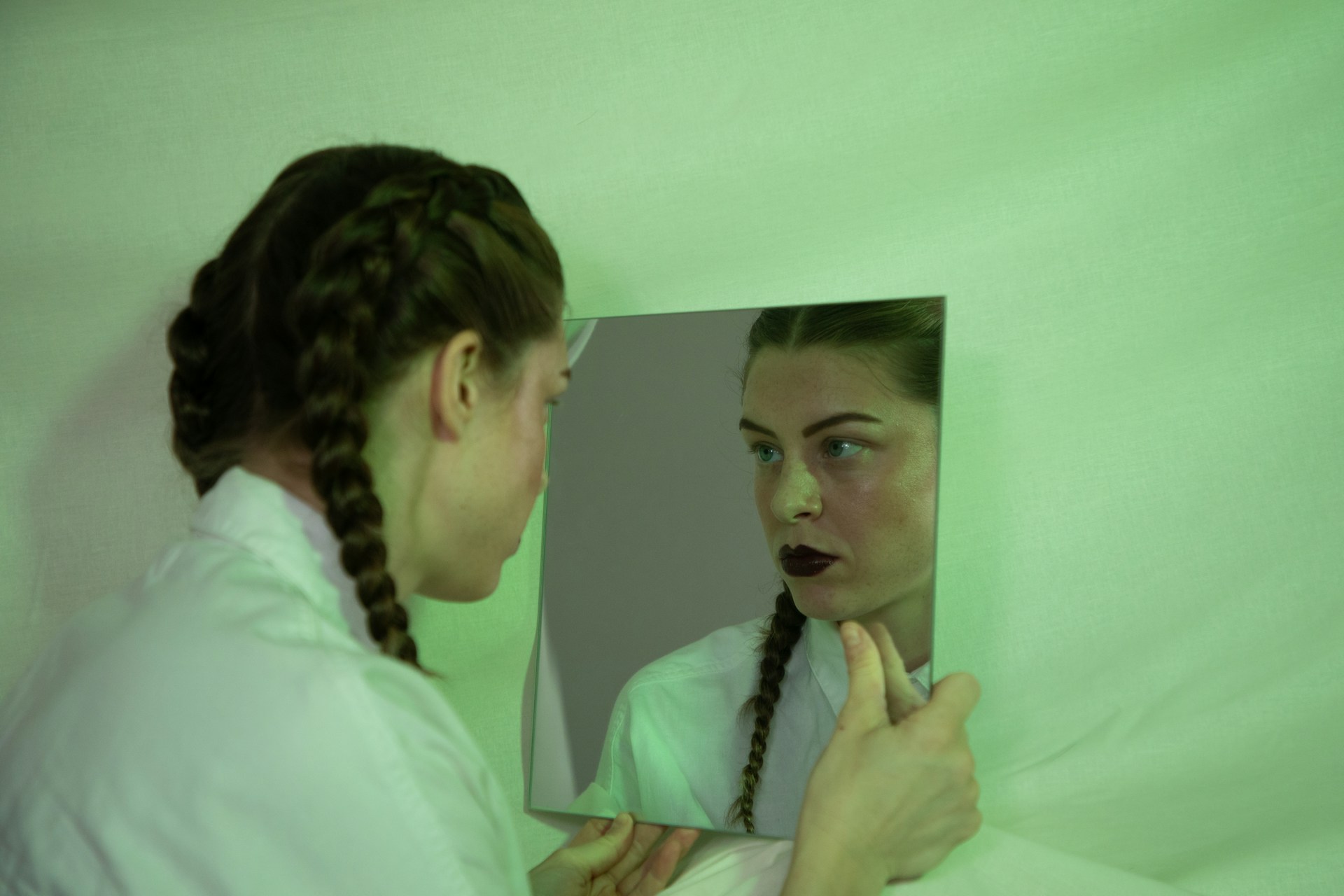 A woman with braided hair and dark lipstick contemplates her reflection in a mirror, her gaze deepening as if exploring the layers of consciousness against a green-tinted background.