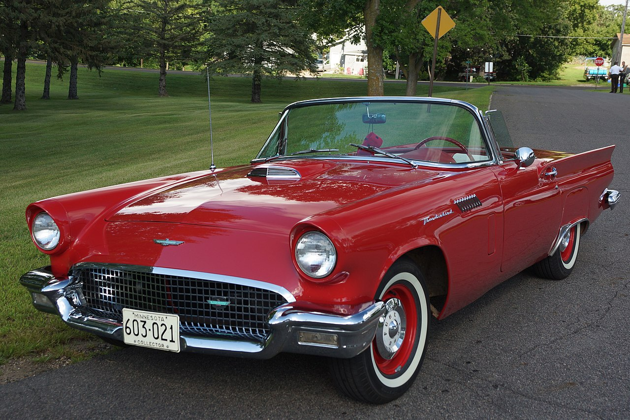 A classic red convertible car parked on a road, surrounded by green grass and trees, with a visible license plate from Minnesota.