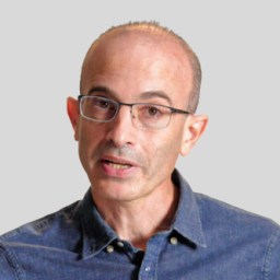 Bald man with glasses wearing a denim shirt speaking against a gray background.