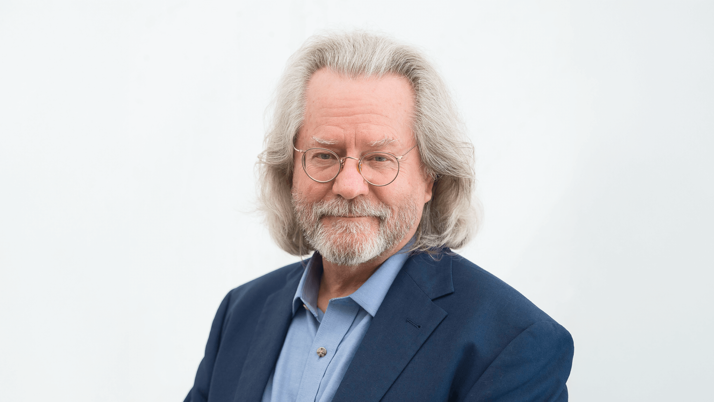 A person with long gray hair, glasses, and a beard, wearing a blue shirt and dark jacket, smiles slightly against a plain white background, exuding a serene aura reminiscent of religious tranquility.