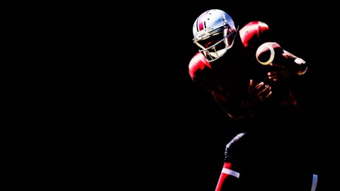 A football player in a red and black uniform, embodying the thrill of dangerous sports, holds a football against a stark black background.