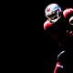 A football player in a red and black uniform, embodying the thrill of dangerous sports, holds a football against a stark black background.