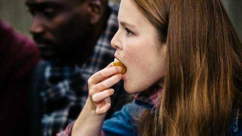 A woman with long hair eats a fruit, holding it near her mouth. A blurred figure is visible in the background.