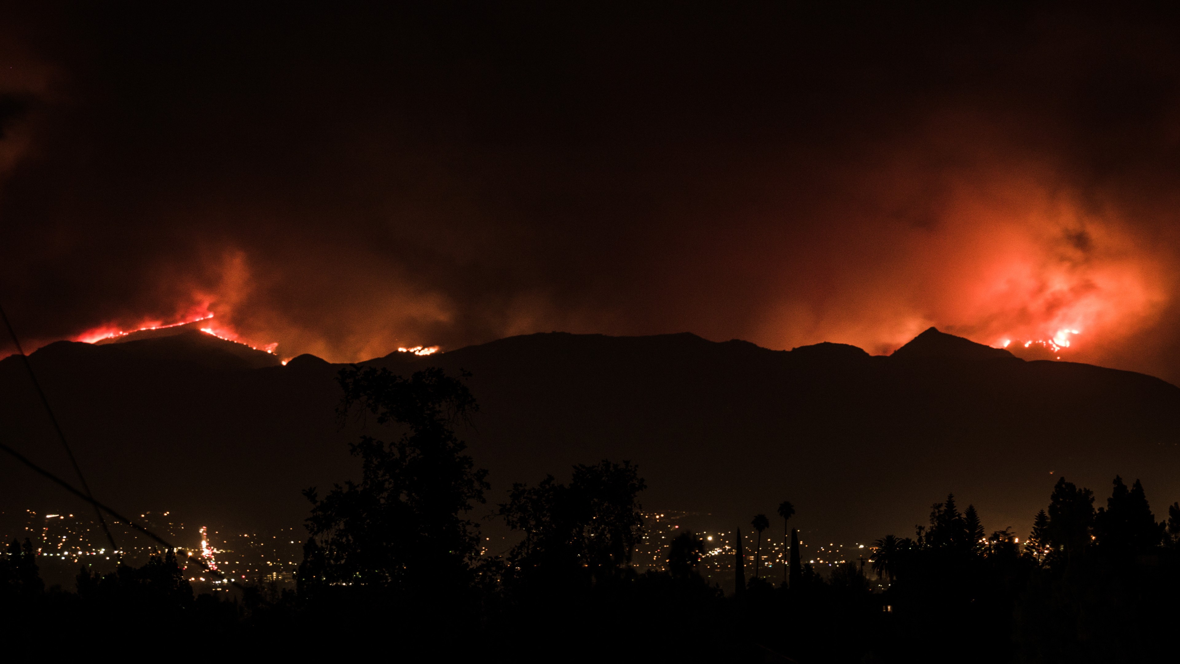 Wildfire burns along a dark mountain ridge at night, with city lights visible below and silhouetted trees in the foreground.