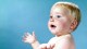 A baby with blonde hair and blue eyes looks upward with a curious expression against a blue background.