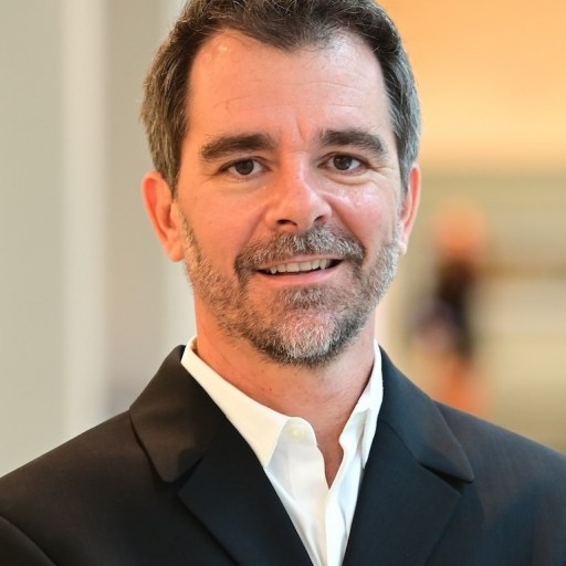 A man with short dark hair and a beard smiles, wearing a black suit jacket and white shirt, standing indoors.