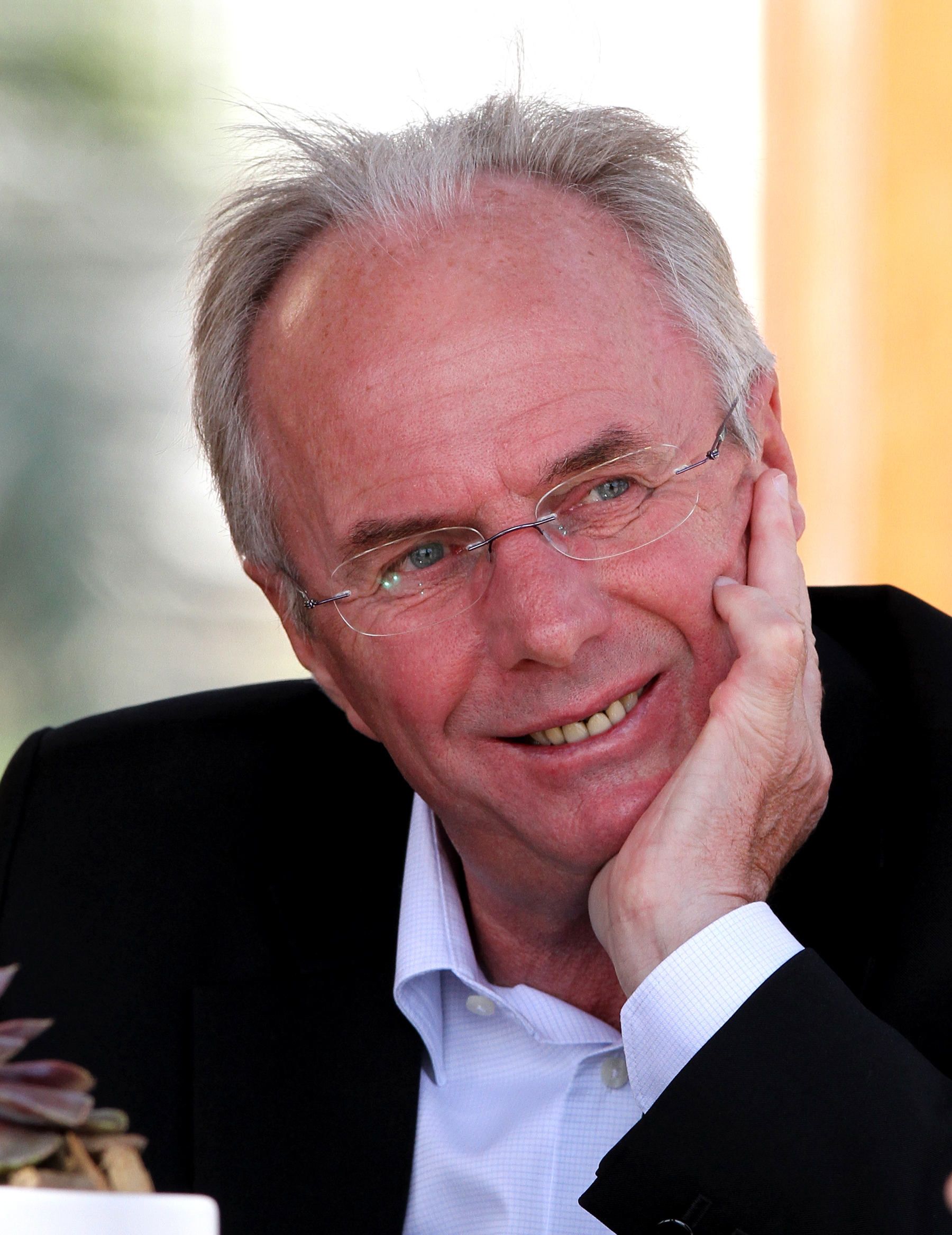 An older man with glasses and a suit smiles while resting his cheek on his hand.