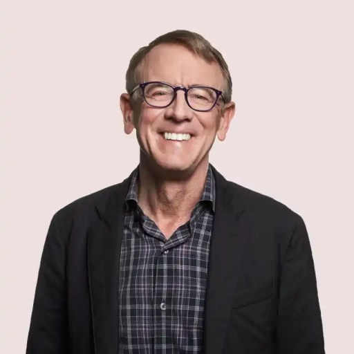 A smiling person with glasses and short hair, wearing a checkered shirt and dark blazer, stands against a plain background.