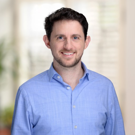 Man with short dark hair and beard wearing a light blue shirt, smiling, standing indoors with a blurred background.