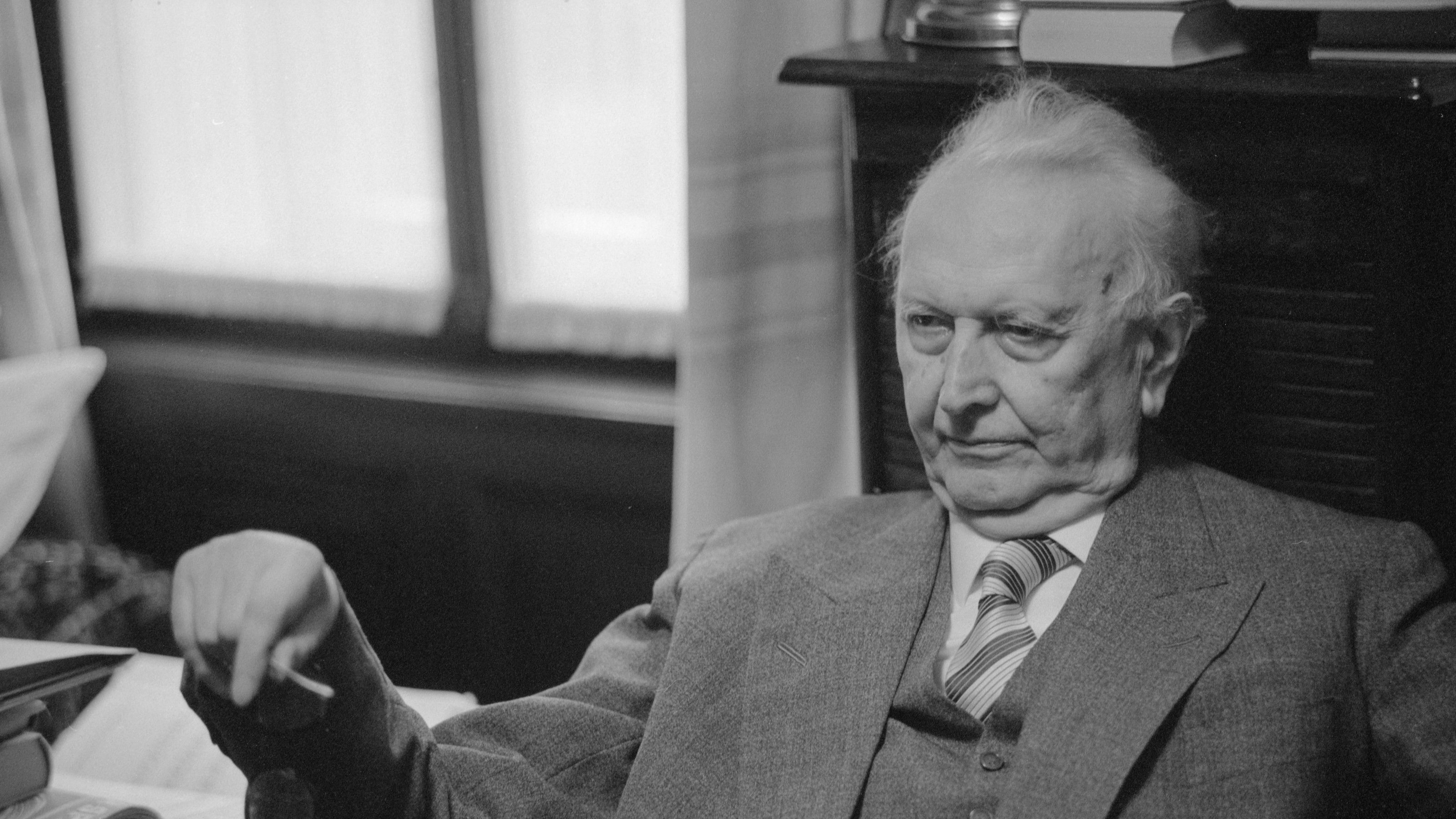 An elderly man in a suit sits indoors, gazing thoughtfully to the side as he contemplates a limit situation. Shelves filled with books and a cabinet form the backdrop of his reflective scene.