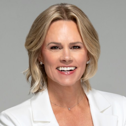 A person with shoulder-length blonde hair, smiling, wearing a white blazer and necklace, against a neutral background.